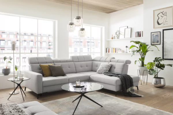 Ecksofa MR4615 aus Feincord in hellgrau mit aufstellbaren Kopfstützen, hellbrauen Zierkissen und einem hellgraugemusterten Teppich mit einem Glascouchtisch