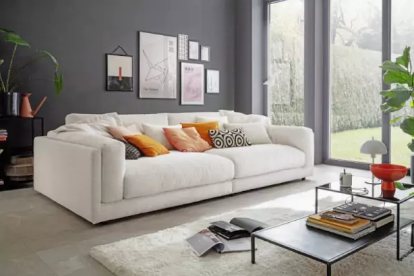 3-Sitzer Cordsofa King Size in beige mit beigen, gelben und gemusterten Kissen. Davor liegt ein hochflorteppich in weiß auf dem ein schwarzer Couchtisch bestehend aus einem tiefen und einem höheren Tisch