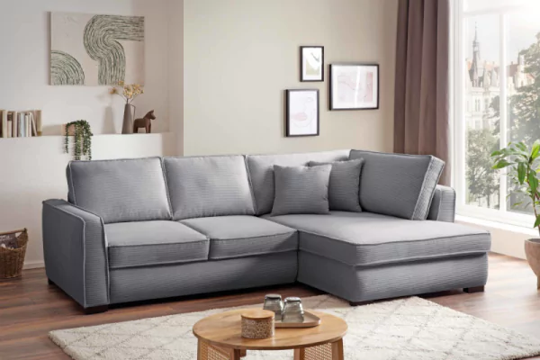 Cordsofa Balou in hellgrau. Das Ecksofa hat zwei hellgraue Zierkissen. Davor steht ein Holzcouchtisch auf einem beigen Teppich mit Rautenmuster