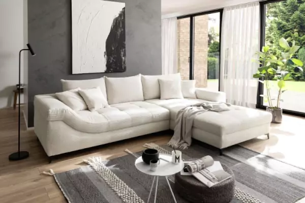 Weiße Eckcouch Perry aus Cord mit farblich passenden Sofakissen. Das Sofa steht auf einem schwarzgemusterten Teppich mit einem weißen Couchtisch und einem schwarzen Hocker